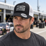 Male model wearing a black-and-white Chevrolet 3D Badge Trucker Cap in a motorsport paddock.